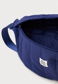 Laukun materiaali on tummansininen tikattu kangas, ja siinä on avattu vetoketju, säädettävä olkahihna sekä valkoinen logotäplä edessä valkoista taustaa vasten.