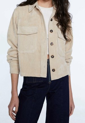 Personne portant une veste boutonnée en velours côtelé beige par-dessus un crop top blanc et un pantalon bleu foncé taille haute avec un détail de fermeture éclair à l'avant.