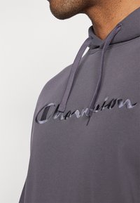 Hoodie gris foncé avec cordon de serrage, mettant en avant un logo "Champion" brodé de manière proéminente avec une finition brillante sur la poitrine.