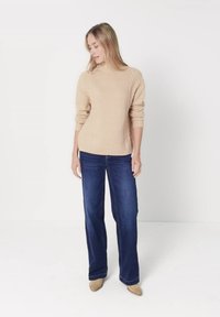 Woman standing, wearing beige cable-knit sweater, dark blue wide-leg jeans, and beige pointed shoes, against plain white background.