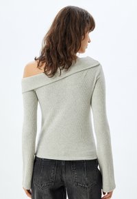Lichtgrijze trui met een off-the-shoulder ontwerp, lange mouwen, geribbelde textuur en een losse pasvorm, gecombineerd met donkere spijkerbroek.