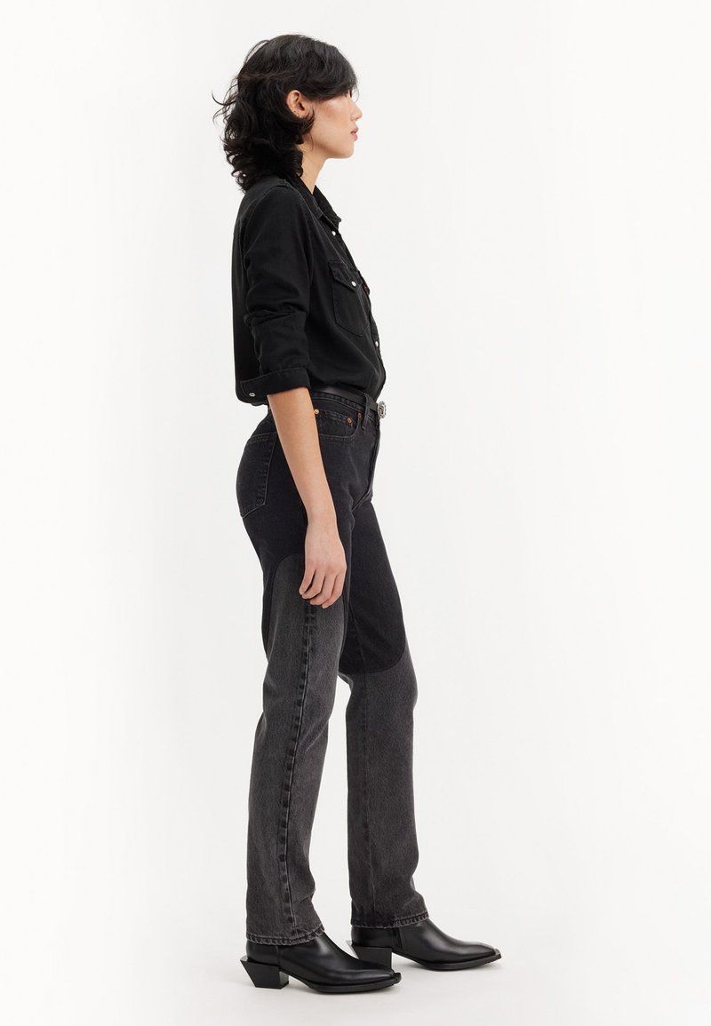 Camisa negra de botones con mangas enrolladas, combinada con pantalones vaqueros gris oscuro con parches en las rodillas y botines negros de punta afilada.