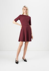 Rochie burgundă cu mânecă scurtă, cu un corset ajustat și fustă evazată. Material neted cu o ușoară strălucire; asociată cu pantofi negri cu vârful ascuțit.