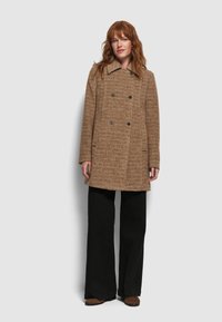 Abrigo marrón texturizado con un diseño de doble botonadura, gran cuello y dos bolsillos frontales. Combinado con pantalones de pierna ancha negros y zapatos estampados.