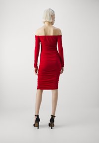 Rochie roșie off-the-shoulder cu mâneci lungi, design pliat și lungime până la genunchi. Modelul poartă pantofi cu toc înalt negri.