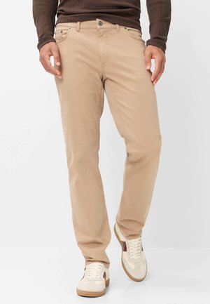 Mand iført beige bukser, hvide sneakers med brune detaljer og en brun langærmet ribtrøje, står mod en lys baggrund.