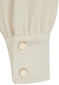 Krém színű, hosszú ujjú mandzsetta két kerek aranygombbal a végén záródik.
