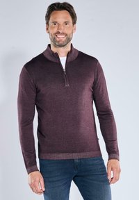 Burgunderfarbener Reißverschluss-Pullover aus Strickstoff mit strukturiertem Finish, ausgestattet mit einem hohen Kragen und langen Ärmeln, kombiniert mit blauen Jeans.