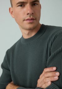 Giovane uomo con i capelli corti che indossa un maglione a coste verde scuro, con le braccia incrociate e lo sguardo leggermente rivolto di lato, su uno sfondo neutro.
