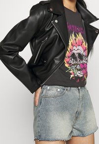 Chaqueta de motorista de cuero negro con cremalleras plateadas, llevada sobre una camiseta gráfica con diseño de calavera y flores. Combinada con pantalones cortos de mezclilla claros.
