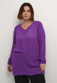 Blouse violette à manches longues avec un col en V, présentant une texture subtile et des fentes sur les côtés, associée à un pantalon noir.