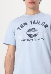 Tricou din bumbac de culoare albastru deschis, cu guler rotund, mâneci scurte și text grafic negru cu un element de design vintage.