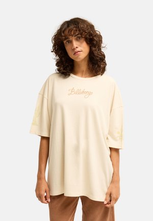 Vrouw met krullend bruin haar, gekleed in een oversized crème Billabong T-shirt en bruine corduroy broek tegen een effen achtergrond.