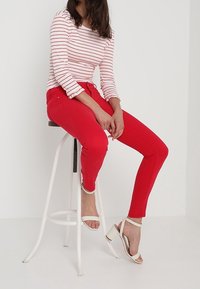 Jean skinny rouge, haut à manches longues rayé avec des lignes rouges, sandales blanches à talons carrés. Design taille haute, silhouette ajustée, tissu lisse.