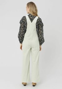 Salopette en velours côtelé vert clair avec une coupe ample, dotée de bretelles ajustables et de deux poches arrière. Porter par-dessus un blouse noire à fleurs.