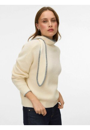 Pull en maille côtelée crème avec un col montant et un long accent drapé gris. Il comporte des manches longues et une coupe décontractée. Porté avec un jean foncé.