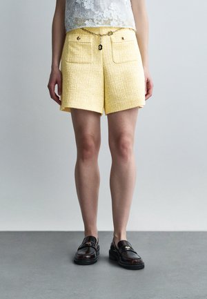 Kobieta w żółtych, fakturowanych szortach z przednimi kieszeniami i łańcuchowym paskiem, białym koronkowym topie oraz czarnych skórzanych mokasynach, stojąca na szarej podłodze.