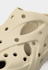 Κοντινή λήψη μπεζ παπουτσιού χωρίς κορδόνια με πολλαπλές οπές αερισμού και ανάγλυφες, σχηματισμένες λεπτομέρειες σχεδίασης.