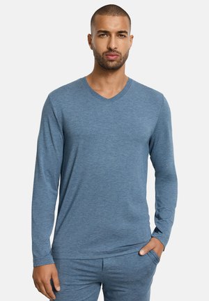 Hombre vistiendo una camisa azul de manga larga con cuello en V y pantalones a juego, de pie con una mano en el bolsillo contra un fondo liso.