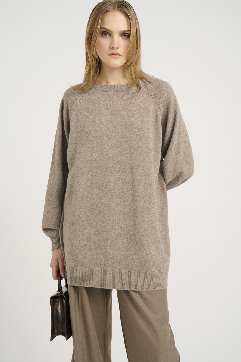 Jasnobrązowy, oversize sweter z raglanowymi rękawami, ściągaczami na mankietach i okrągłym dekoltem, zestawiony z szerokimi, beżowymi spodniami i małą torebką.