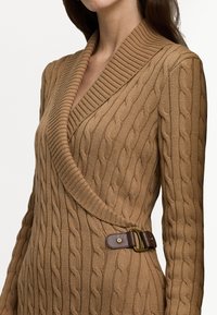 Svetr s kabelovým pletením v béžové barvě s hlubokým výstřihem do V, širokým ribbed límcem a hnědým koženým sponou na boku. Měkká textura.
