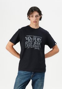 Black cotton t-shirt featuring a vintage graphic print in white and grey, crew neckline, and short sleeves. Worn with denim jeans.