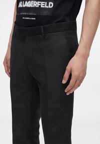 Gros plan d'une personne portant un pantalon noir sur mesure et un t-shirt noir Karl Lagerfeld avec un texte visible sur la poitrine.