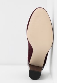 Semelle d'une chaussure à talon haut en daim bordeaux avec un dessous beige lisse et un embout de talon noir texturé sur fond blanc.