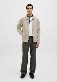 Beige ritsjack met een kraag, gecombineerd met een witte trui en donkergrijze geplooide broek, aangevuld met een blauwe patroon sjaal en zwarte schoenen.