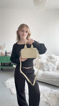 Νεαρή γυναίκα με μαύρο σύνολο κρατάει μία μπεζ τσάντα Calvin Klein, στέκεται σε φωτεινό σαλόνι με καναπέ, μαξιλάρια και πράσινη ντουλάπα.