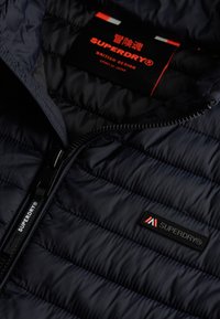 Navy puffer jacket with quilted texture, featuring a black label and contrasting logo. Zipper closure with branded pull tab.