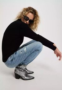 Silberne Ankle-Boots mit spitzer Zehe und Blockabsatz, kombiniert mit hellblauen, verwaschenen Jeans und einem schwarzen Langarmoberteil. Lockige Haare und Sonnenbrille.