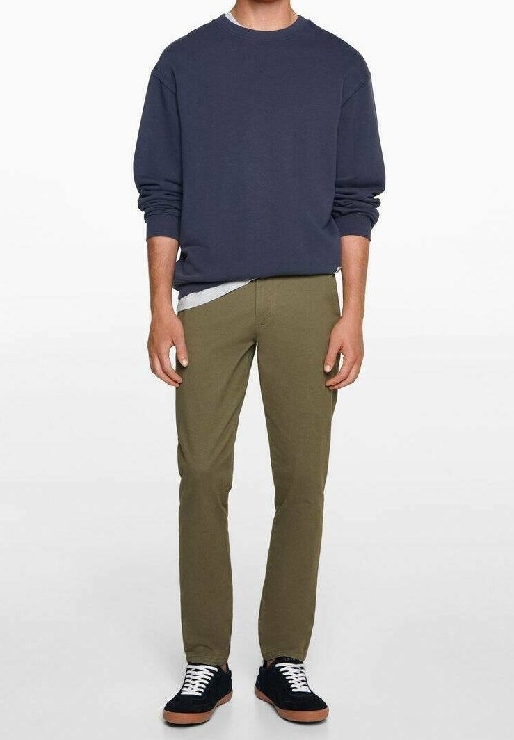 Homme portant un sweat-shirt bleu marine, un pantalon slim olive et des baskets noires à lacets blancs, debout devant un fond uni.