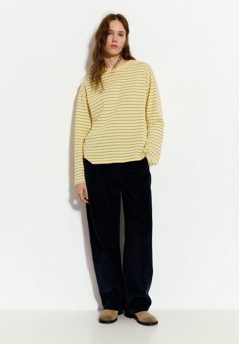 Geel en marine gestreepte oversized trui gecombineerd met losse marineblauwe corduroy broek; bruine suède enkellaarsjes maken de outfit af.