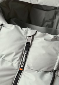 Lys grå dunjakke med fuld frontlynlås, Superdry branding og tekstureret stof med en blød inderside.