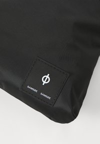 Svart stoffpose med en teksturert overflate og et rektangulært merke med en hvit logo og merkenavnet "SAMSØE SAMSØE" i en stilren skrifttype.