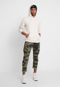 Hettegenser i krem med kengurulomme, kombinert med camouflage cargo-bukser med flere lommer og oppbrettede mansjetter, fullført med hvite joggesko.