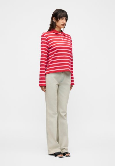 Γυναίκα όρθια φορώντας κόκκινη και ροζ ριγέ μακρυμάνικη πόλο μπλούζα, ανοιχτό μπεζ παντελόνι και μαύρα ανοιχτά σανδάλια σε λευκό φόντο.