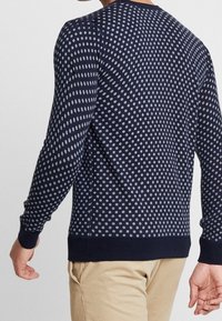 Muž v tmavě modrém svetru s světle šedými puntíky, v béžových kalhotách, zobrazen zezadu a mírně se otáčí doleva.