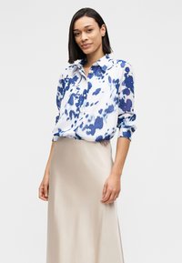 Πουκάμισο με κουμπιά σε λευκό χρώμα με μπλε αφηρημένο σχέδιο, διαθέτει κλασικό γιακά και μακριά μανίκια. Φορεμένο με μπεζ σατέν φούστα.