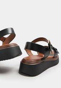 Svarta lädersandaler med plattform, två breda remmar, en rosettdetalj och en brun lädersula. Har en texturerad, tjock sula för stöd.