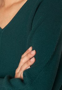 Suéter de punto color teal con cuello en V, que presenta un patrón de canalé texturizado. Las mangas tienen un corte holgado, acentuado por un puño doblado de estilo informal.