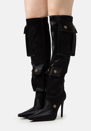 Bottes hautes noires en cuir atteignant le genou, avec un bout pointu, des poches décoratives, des accents en métal doré et des talons aiguilles. Texture lisse sur toute la surface.