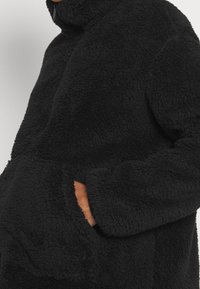 Schwarze Fleecejacke mit hohem Kragen, Reißverschluss und großen Fronttaschen. Weicher, strukturierten Stoff mit lockerer Passform.