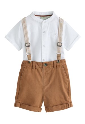 Junge lächelnd, trägt ein weißes kurzärmeliges Hemd mit Knöpfen, braune Shorts mit Hosenträgern, weiße Socken und weiße Turnschuhe.