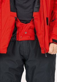 Rote wasserdichte Jacke mit schwarzem Innenfutter, ausgestattet mit Druckknopfverschluss und verstellbaren Manschetten, kombiniert mit schwarzen, strukturierten Hosen.