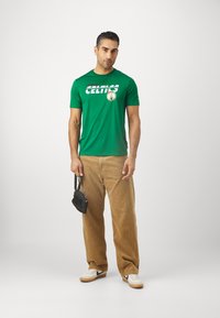 Zöld Boston Celtics témájú póló, merész fehér betűkkel, bézs kordbársony nadrággal és fehér tornacipővel kombinálva, egy kis fekete táskát tartva.