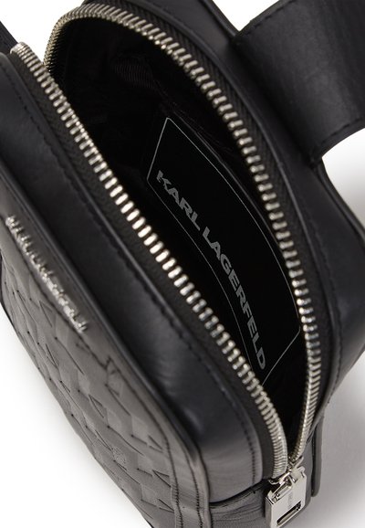 Sac en cuir noir avec un motif tissé texturé, fermeture éclair argentée et étiquette intérieure. Forme rectangulaire compacte avec une sangle durable.