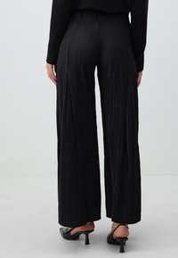 Pantalons larges noirs plissés avec une taille haute, présentant des plis verticaux et une texture lisse, associés à des talons ouverts noirs.