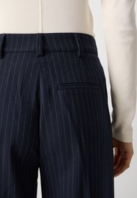 Pantaloni navy a righe sottili con finitura liscia, dotati di passanti per cintura e una tasca a filetto sul lato destro. Presentati con una maglietta chiara.